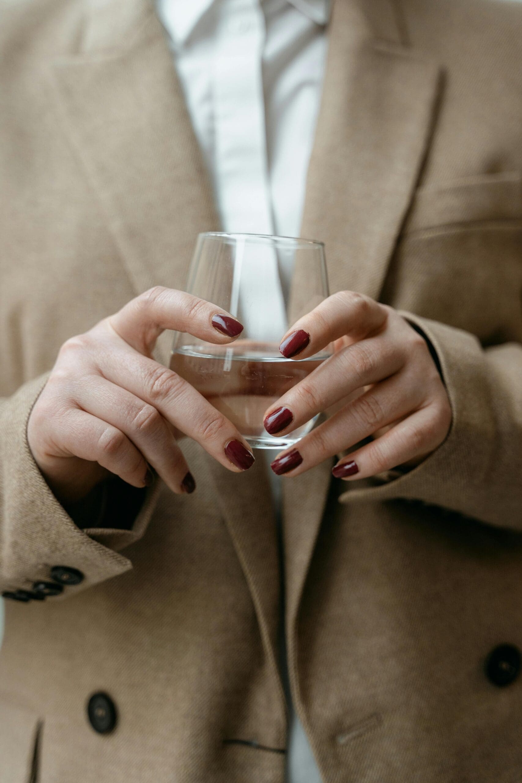 Close-up of a woman holding a glass of water, wearing a beige coat and white shirt, symbolizing wellness and hormone balance.